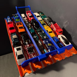 May include: A blue plastic multi-lane track organizer holding a collection of various die-cast toy cars in red, orange, black, white, green, yellow, and blue. The track features five parallel slots, each containing multiple small toy vehicles.