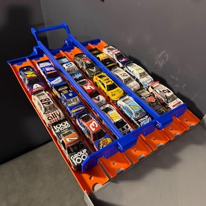 May include: A toy race track with multiple miniature race cars. The track is orange with blue launch mechanisms. The cars are brightly colored with various sponsor logos and numbers, including "AXALTA", "GROUP 100", and "RVSTOP".