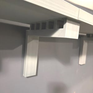 Op de afbeelding: Een witte, geometrische plankbeugel, gemonteerd op een grijze muur. De beugel heeft een rechthoekig ontwerp met een reeks verticale steunen en een onderste, L-vormige steun. De plank is bevestigd aan een horizontaal oppervlak.