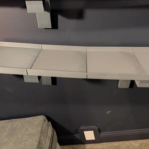 May include: A gray, modular wall-mounted track system for toy cars. The track is composed of several gray rectangular sections supported by gray brackets. The track is mounted on a dark blue wall. Orange track pieces are visible on the left.