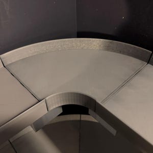 May include: A modular grey plastic corner shelf component with a smooth, flat surface and a raised outer edge. The piece features a curved inner cutout and straight side edges designed to connect with other similar segments to form a larger unit.