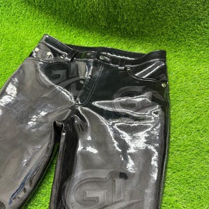 May include: Black, high-waisted, shiny pants with a glossy finish. The pants have a classic five-pocket design and a button and zipper closure. The material reflects light, creating a sleek, modern look. The pants are shown against a green background.