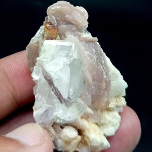 May include: A raw mineral specimen featuring a cluster of translucent, pale blue-green crystals embedded within a matrix of layered, light pinkish-tan lepidolite and white feldspar, with a small, distinct amber-colored crystal at the top.