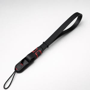 May include: A black wrist lanyard featuring a flat strap with a loop at one end and a thin cord attachment at the other. The connector piece is black with red accents and a circular red detail, providing a secure way to carry small electronic devices.