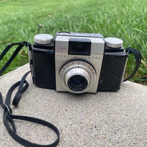 May include: A vintage Kodak Pony II camera with a black leatherette body and silver faceplate. The camera has a black strap and two silver dials on top. The lens is surrounded by a red and silver dial. The camera is on a concrete surface.