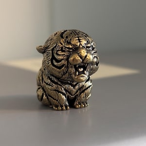 May include: A small, metallic, brass-coloured figurine of a seated tiger. The tiger has a rounded, stylised body with deeply etched black stripes and a detailed face with an open mouth, bared teeth, and focused eyes, capturing a fierce expression.