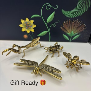 May include: A set of brass insect ornaments, featuring a dragonfly, bee, grasshopper, and praying mantis. The text "Gift Ready" is visible with a gift box icon. The insects are displayed on a white surface with a floral design in the background.