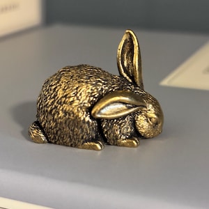 May include: A small, antique-style brass figurine of a crouching rabbit. The metal sculpture features a textured, fur-like finish across its body, with one ear standing upright and the other folded down against its head in a resting position.