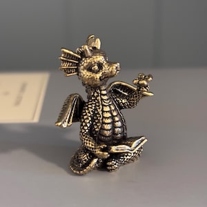 May include: A small, antique brass-coloured metal figurine of a seated dragon. The dragon has textured scales, small wings, and horns, and it is holding an open book in its lap while gesturing with one clawed hand towards a small flower.