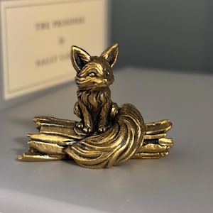 May include: A small, metallic, brass-toned figurine of a fox sitting atop a textured log. The fox has pointed ears, detailed fur texture, and a long, curled tail that wraps around the base of the log. The object has a weathered, antique finish.