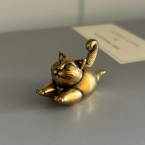 May include: A small, metallic gold-toned cat figurine in a playful, stretching pose. The cat has a rounded body, pointed ears, and a textured, upright tail. Its eyes are closed in a contented expression, and its front paws are extended forward.