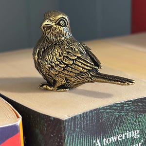 May include: A gold-coloured bird ornament with detailed feathering and a focused expression. The bird is positioned on a book, highlighting its intricate design and metallic finish. The figurine's pose suggests a watchful presence.