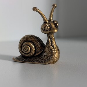 May include: A small, metallic bronze-coloured snail figurine with a textured surface. The snail features a coiled shell, two prominent eye stalks with rounded tips, and large, expressive eyes. The overall finish has a weathered, antique appearance.