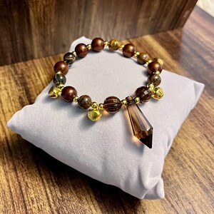 Peut inclure: Un bracelet de perles composé de perles marron et œil-de-tigre alternées, de petites entretoises en métal doré et de trois petites clochettes dorées. Un pendentif facetté en forme de goutte marron translucide pend au centre.