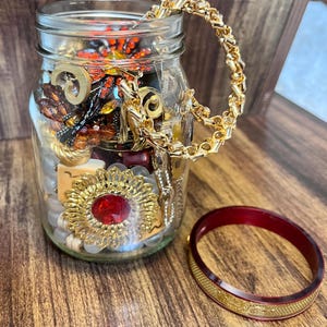 May include: A clear glass jar filled with assorted costume jewellery, including a large gold-tone brooch with a central red stone, a gold chain bracelet draped over the rim, and various beads. A separate dark red and gold-tone bangle rests nearby.
