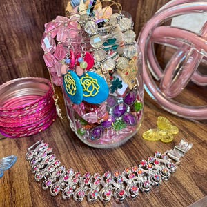 May include: A clear glass jar filled with assorted jewellery including pink faceted beads, turquoise earrings with yellow rose designs, and various charms. A silver-tone bracelet with iridescent stones lies in front, alongside a stack of pink bangles.