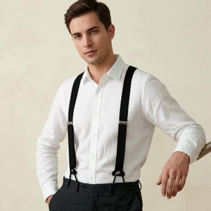 May include: A crisp white long-sleeved button-down shirt paired with solid black suspenders featuring silver-toned metal hardware clips. The suspenders are worn over the shirt, which is tucked into dark trousers, creating a clean and structured look.
