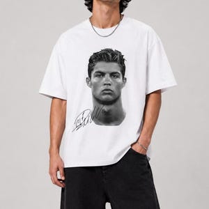 Peut inclure: Un t-shirt blanc à manches courtes avec un portrait photographique en noir et blanc d'un homme aux cheveux coiffés sur la poitrine. Sous le portrait se trouve une signature noire manuscrite. Le t-shirt a un col rond et une coupe ample.
