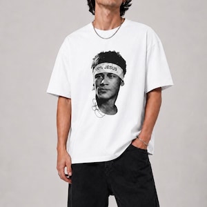 Peut inclure: Un t-shirt blanc à manches courtes avec une impression graphique en noir et blanc représentant une personne portant un bandeau sur lequel est écrit 100% JESUS. Le motif inclut une signature stylisée sous le portrait de l'individu.