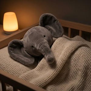 K&ouml;nnte beinhalten: Ein graues Pl&uuml;schelefanten-Spielzeug liegt auf einer cremefarbenen Strickdecke in einem Holzbettchen. Eine kleine, leuchtende Lampe steht auf einem Nachttisch im Hintergrund, mit einem Korb voller Teddyb&auml;ren.
