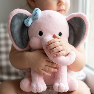 K&ouml;nnte beinhalten: Ein rosa Pl&uuml;schelefant mit grauen Ohren und einer hellblauen Schleife. Der Elefant hat schwarze Augen. Das Spielzeug ist weich und kuschelig, ideal f&uuml;r ein Baby oder Kleinkind.