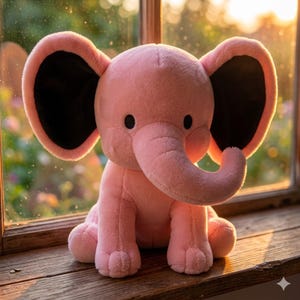 K&ouml;nnte beinhalten: Ein rosa Pl&uuml;schelefant mit gro&szlig;en, schwarz umrandeten Ohren, einem R&uuml;ssel und kleinen schwarzen Augen. Das Stofftier sitzt auf einer Holzoberfl&auml;che in der N&auml;he eines Fensters und wirkt kuschelig.