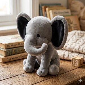 K&ouml;nnte beinhalten: Ein graues Pl&uuml;schelefant-Spielzeug mit schwarz gef&uuml;tterten Ohren sitzt auf einer Holzoberfl&auml;che. Der Elefant hat einen R&uuml;ssel, kleine schwarze Augen und sitzt. Im Hintergrund sind B&uuml;cher und ein Holzblock zu sehen.