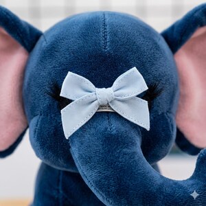 K&ouml;nnte beinhalten: Ein blaues Pl&uuml;sch-Elefanten-Spielzeug mit einer hellblauen Schleife am R&uuml;ssel. Der Elefant hat rosa Innenohren und schwarze Wimpern. Das Spielzeug ist aus weichem, samtigem Material gefertigt.