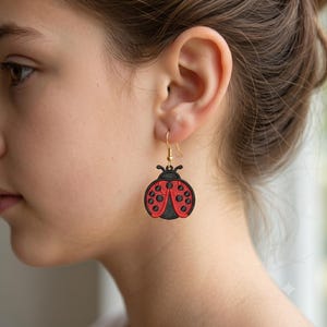 Puede incluir: Un pendiente colgante con un diseño circular de mariquita que presenta un cuerpo negro texturizado y alas rojas vibrantes adornadas con puntos negros. El dije de mariquita está suspendido de un sencillo gancho de metal en tono dorado.