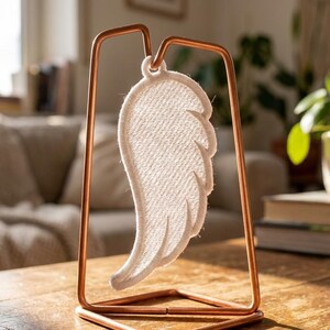 Puede incluir: Un ala de &aacute;ngel decorativa de tela blanca, suspendida dentro de un marco de metal color cobre. El ala presenta costuras detalladas y se exhibe sobre una superficie de madera, creando una exhibici&oacute;n serena y art&iacute;stica.