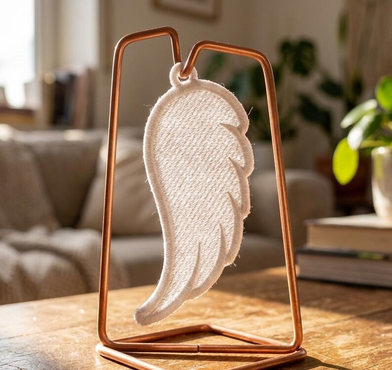 Puede incluir: Un ala de &aacute;ngel decorativa de tela blanca, suspendida dentro de un marco de metal color cobre. El ala presenta costuras detalladas y se exhibe sobre una superficie de madera, creando una exhibici&oacute;n serena y art&iacute;stica.