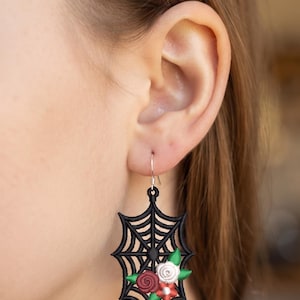 May include: A black embroidered spider web earring featuring a cluster of small fabric flowers in shades of red, white, and pink, accented with green leaves. The earring hangs from a silver-toned metal hook and has a pointed, geometric shape.