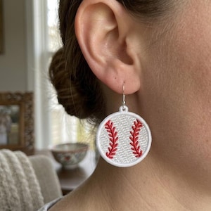 May include: A circular earring featuring a white textured background with red embroidered stitching that mimics the appearance of a baseball. The design is enclosed in a white stitched border and hangs from a silver-toned metal hook.
