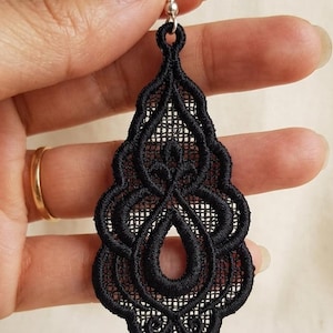 Pode incluir: Um brinco de renda preta com um gancho prateado. O brinco tem um design ornamentado e vazado em forma de gota. O padrão intrincado é visível contra um fundo claro.