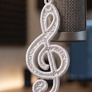 May include: A white embroidered treble clef ornament featuring a detailed stitched texture and a small loop at the top for hanging, suspended in front of a silver studio microphone with a metallic mesh grille and a cylindrical body.