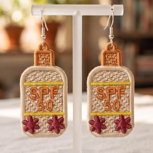May include: A pair of embroidered earrings shaped like sunscreen bottles. The beige textured fabric features the orange text "SPF 50" above two dark red flower shapes. The bottle caps are orange, and the earrings hang from silver-toned metal hooks.