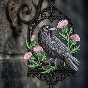 May include: A black embroidered wall hanging featuring a detailed grey raven perched on a branch within a gothic-style frame. The design includes vibrant green stems and pink thistle flowers surrounding the bird, all crafted with intricate thread work.