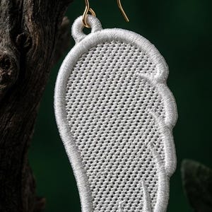 Puede incluir: Un pendiente blanco bordado en forma de ala con un gancho dorado. El pendiente est&aacute; hecho de hilo blanco y tiene una superficie detallada y texturizada. Cuelga de un trozo de madera sobre un fondo verde oscuro.