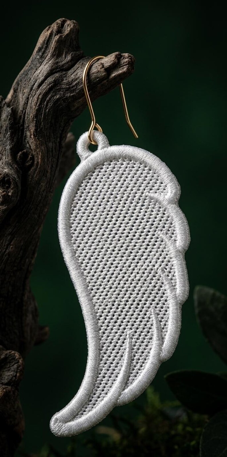 Puede incluir: Un pendiente blanco bordado en forma de ala con un gancho dorado. El pendiente est&aacute; hecho de hilo blanco y tiene una superficie detallada y texturizada. Cuelga de un trozo de madera sobre un fondo verde oscuro.