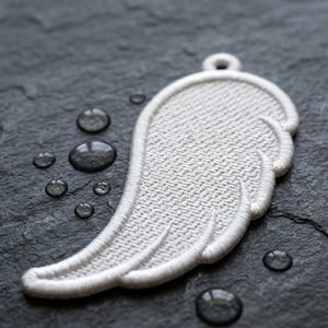 Puede incluir: Un parche blanco bordado en forma de ala, con un lazo en la parte superior para colgarlo. El ala tiene una superficie texturizada y un borde blanco. El fondo es una superficie texturizada gris oscuro con gotas de agua.