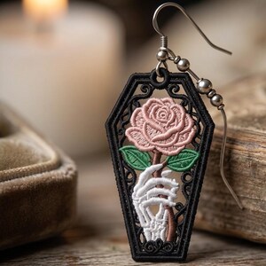 May include: An embroidered earring featuring a black coffin-shaped frame containing a pink rose held by a white skeletal hand. The design is suspended from a silver-toned metal hook with small spherical beads, showcasing detailed needlework.