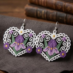 May include: A pair of heart-shaped lace earrings featuring intricate white scrollwork borders. The center of each heart displays an embroidered purple iris flower with green leaves, accented by small white daisies and purple blossoms with yellow centers.