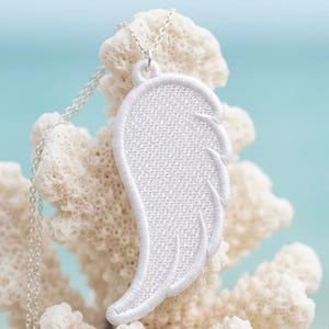 Puede incluir: Collar con colgante de ala bordada en blanco y fina cadena plateada. El colgante de ala est&aacute; detallado con l&iacute;neas cosidas, y el collar se muestra sobre un fondo de coral y un suave azul.