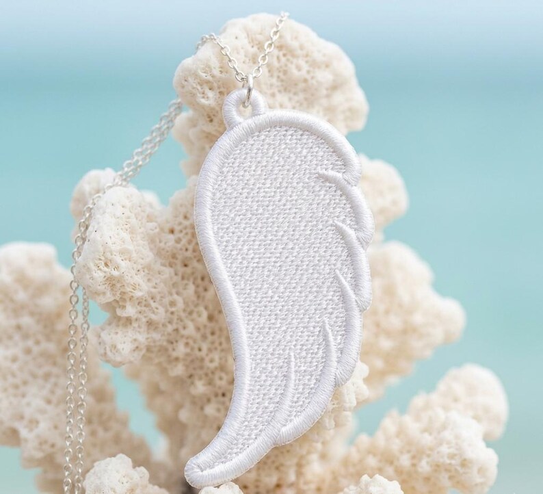Puede incluir: Collar con colgante de ala bordada en blanco y fina cadena plateada. El colgante de ala est&aacute; detallado con l&iacute;neas cosidas, y el collar se muestra sobre un fondo de coral y un suave azul.