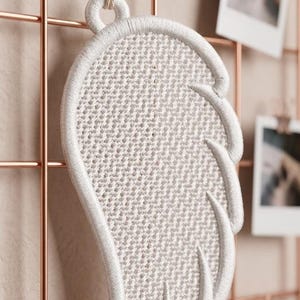 Puede incluir: Un adorno de pared blanco bordado con forma de ala. El ala est&aacute; hecha de un material tejido y est&aacute; sujeta a una rejilla de color cobre. Peque&ntilde;as fotos est&aacute;n sujetas a la rejilla. El ala est&aacute; suspendida por una cuerda de color natural.
