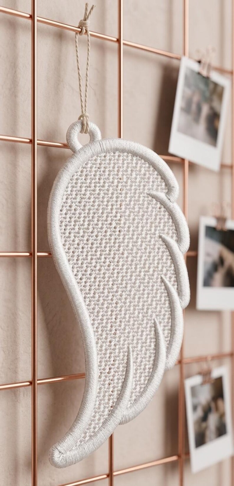 Puede incluir: Un adorno de pared blanco bordado con forma de ala. El ala est&aacute; hecha de un material tejido y est&aacute; sujeta a una rejilla de color cobre. Peque&ntilde;as fotos est&aacute;n sujetas a la rejilla. El ala est&aacute; suspendida por una cuerda de color natural.