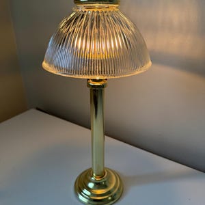 Puede incluir: Una pequeña lámpara de mesa con una pantalla de vidrio transparente y acanalado y una base y un tallo de metal dorado. La lámpara está encendida, emitiendo un brillo cálido. La base es redonda y decorativa.