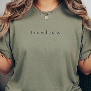May include: Olive green t-shirt with the phrase "this will pass" printed in black. The shirt has short sleeves and a crew neck. The model is wearing blue jeans and a gold necklace.