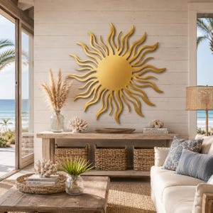 May include: A large, circular gold sun wall sculpture with wavy, flame-like rays extending from a central disc. The metal piece is mounted on a white horizontal wood-paneled wall above a wooden console table in a coastal-themed living space.