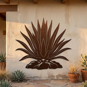 May include: A rust-coloured metal wall art piece shaped like an agave plant with sharp, pointed leaves radiating from a central base. The cutout design features a textured, weathered finish and is mounted flat against a light-coloured stucco wall.